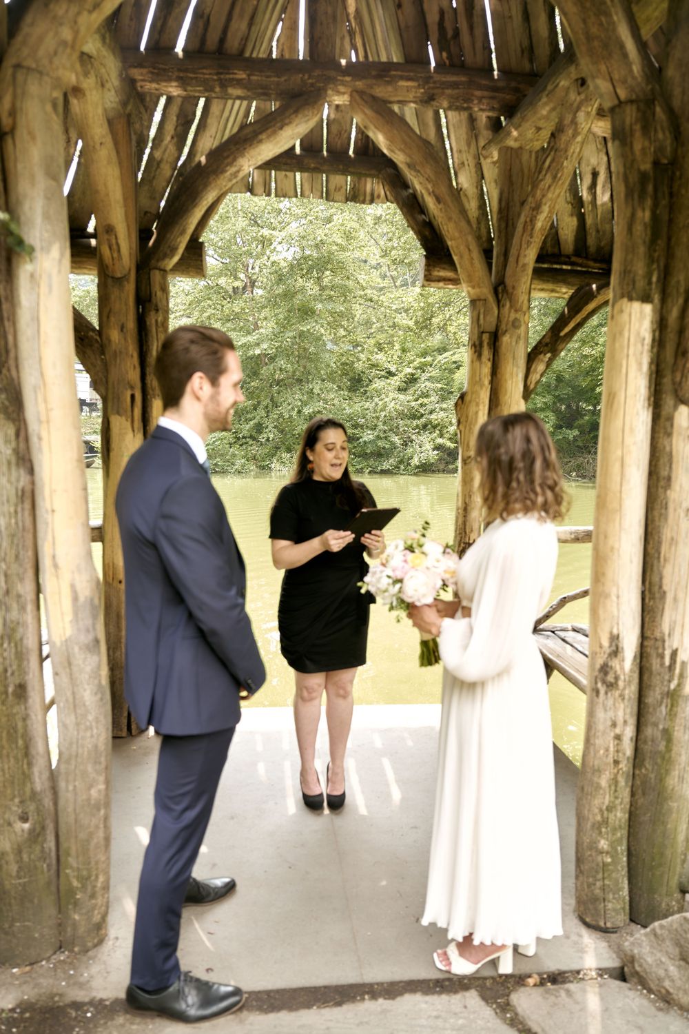 A small wedding ceremony takes place under a rustic wooden gazebo surrounded by greenery. Wedding photoshoot