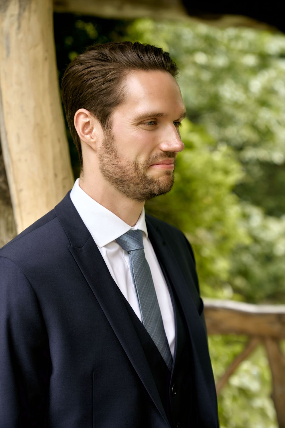 A groom in a dark suit and tie looks contemplatively to the side while standing outdoors with greenery in background. Wedding photoshoot