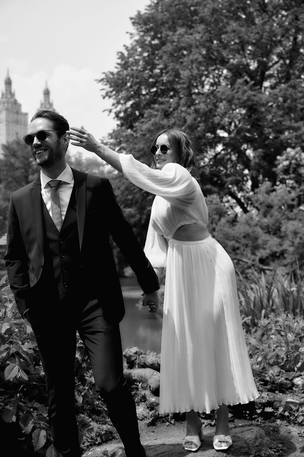 A playful black and white wedding photo of a couple in a garden with city architecture visible behind trees. Wedding photoshoot
