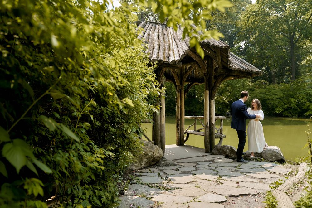 A couple shares an intimate moment under a wooden gazebo surrounded by lush greenery on a stone pathway. Wedding photoshoot