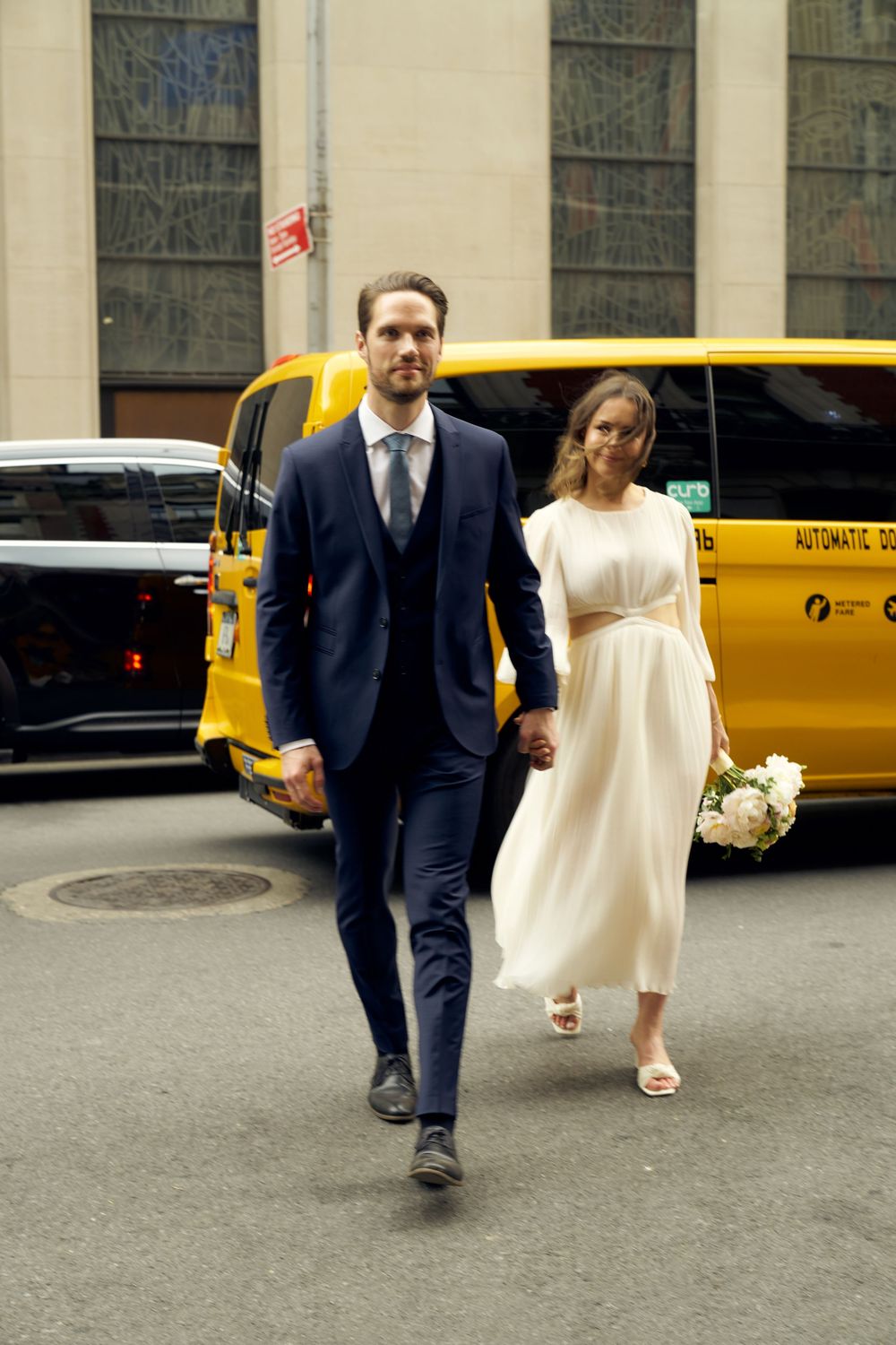 A bride in a white dress and groom in navy suit walk along a city street with yellow taxis in the background. Wedding photoshoot