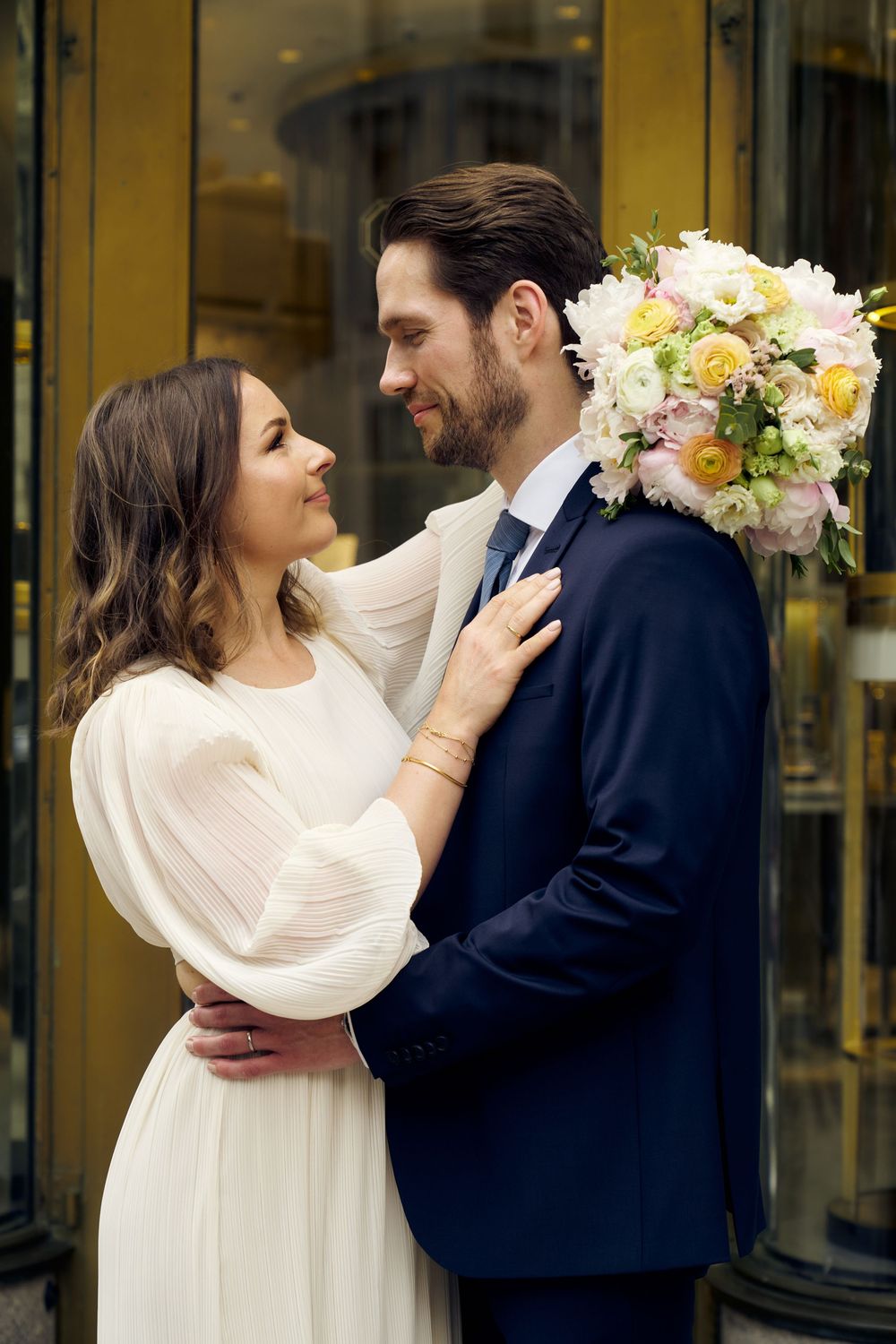 A romantic wedding couple embraces outdoors in front of golden doors, with the groom holding a soft pink and white bouquet. Wedding photosho