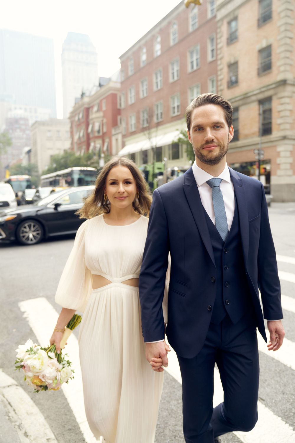 A bride in a flowing cream dress walks hand in hand with a groom in a dark suit along a city street. Wedding photoshoot
