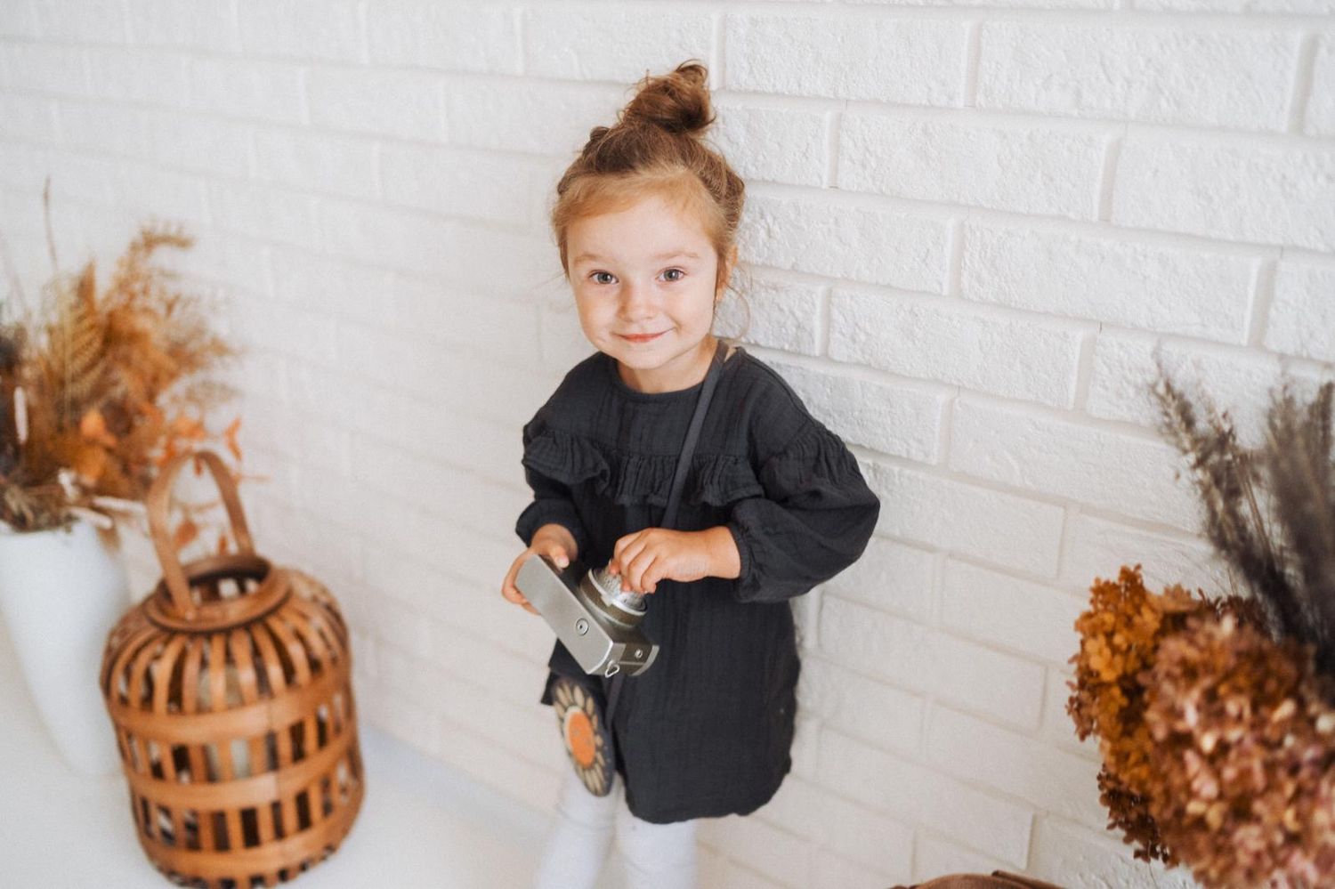 Child in black outfit posing against white brick wall with fall decorations.
