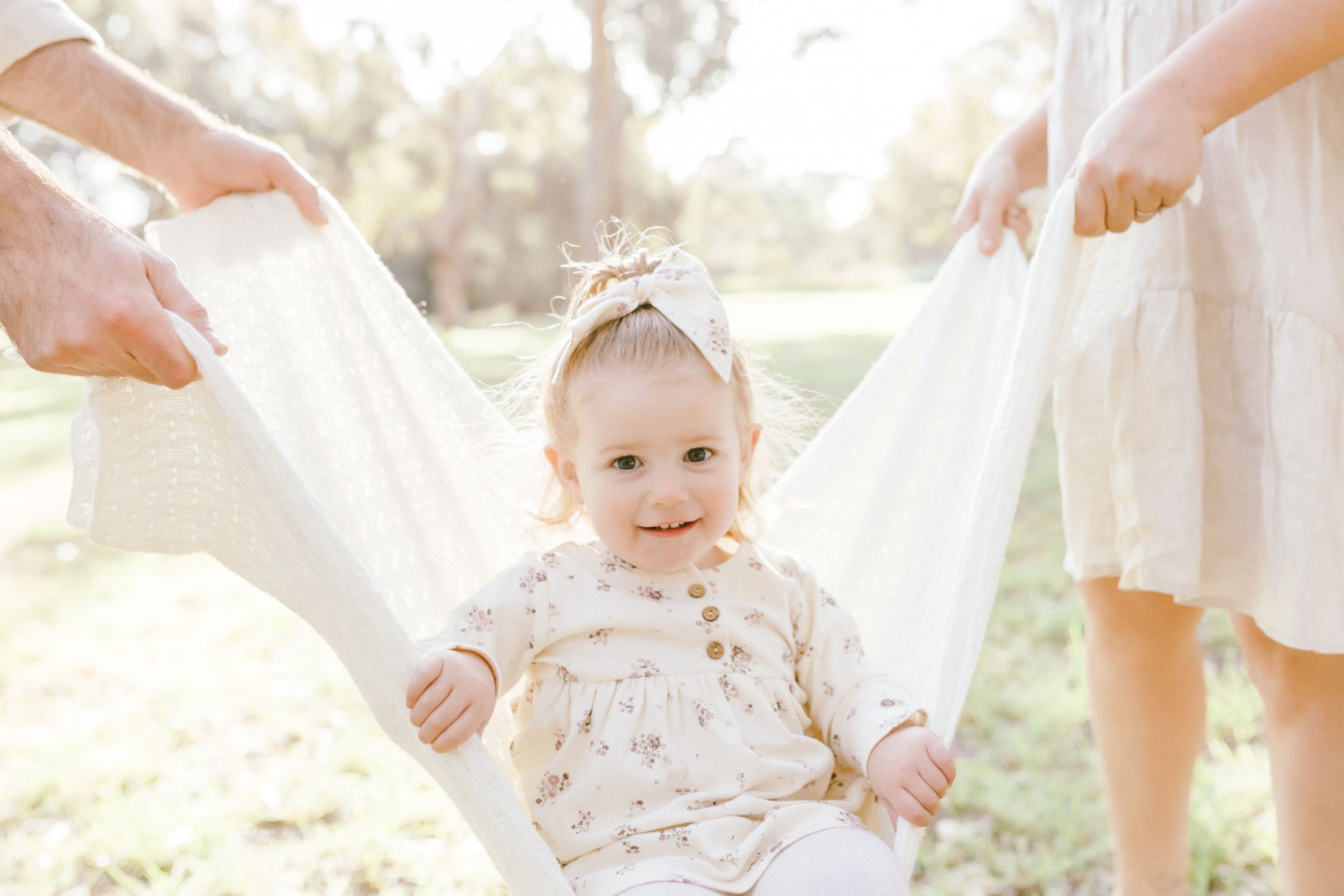 A child in a white lace dress plays outdoors in soft sunlight while standing amongst trees and greenery.