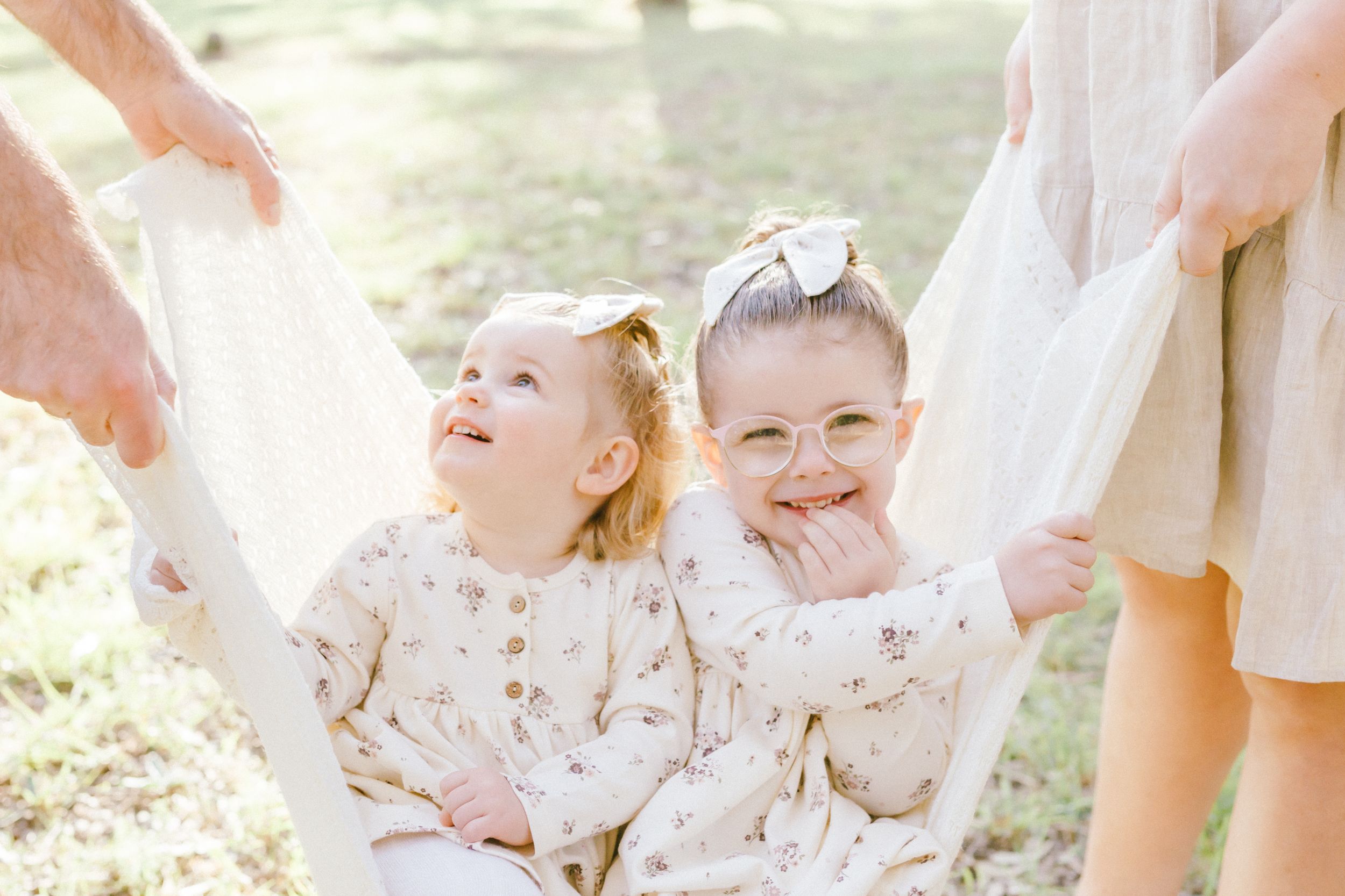 A happy family in coordinated white and beige outfits poses together outdoors in bright sunlight.