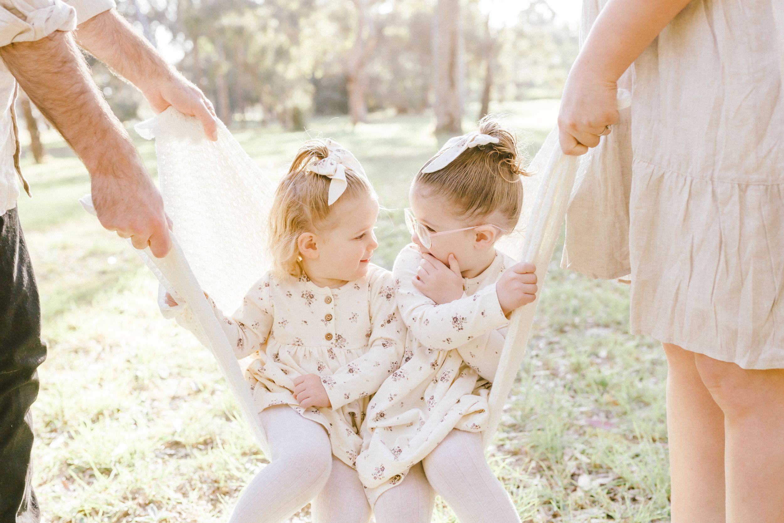 Two toddlers in floral dresses and white leggings laugh together in a sunlit outdoor setting.