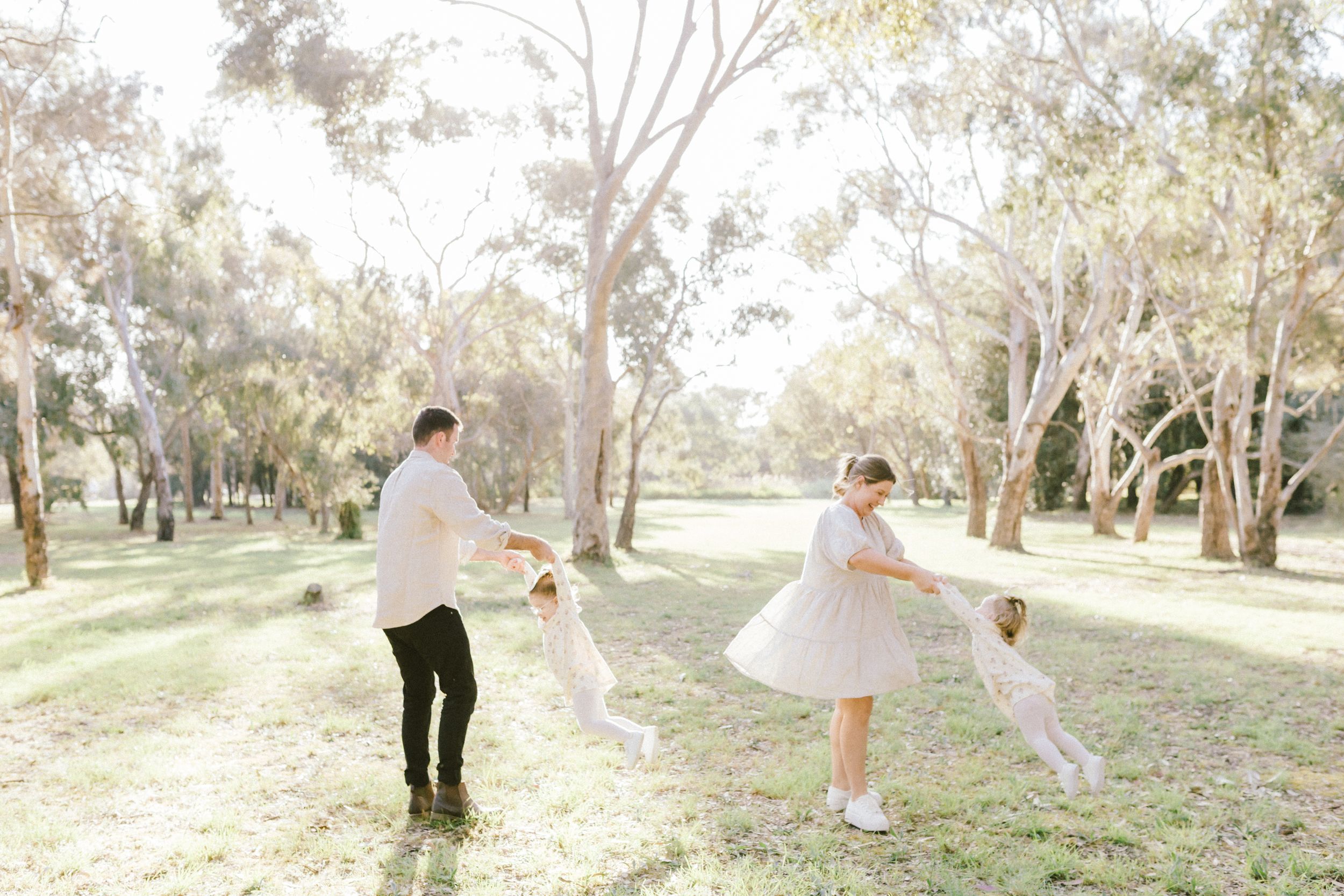 A couple in wedding attire playfully dances and spins together in a sunlit outdoor park setting.