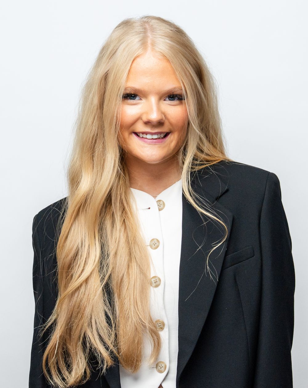 Professional portrait of someone wearing a black blazer and white button-up shirt with blonde hair against a light background.
