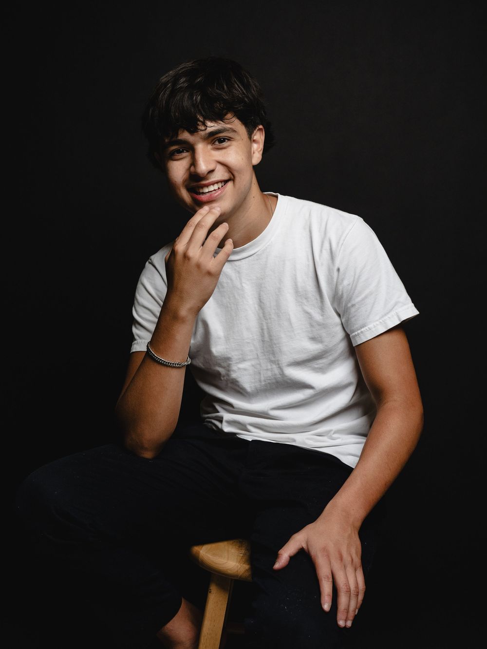 Person in white t-shirt sitting on wooden stool against dark background with natural smile and relaxed pose.