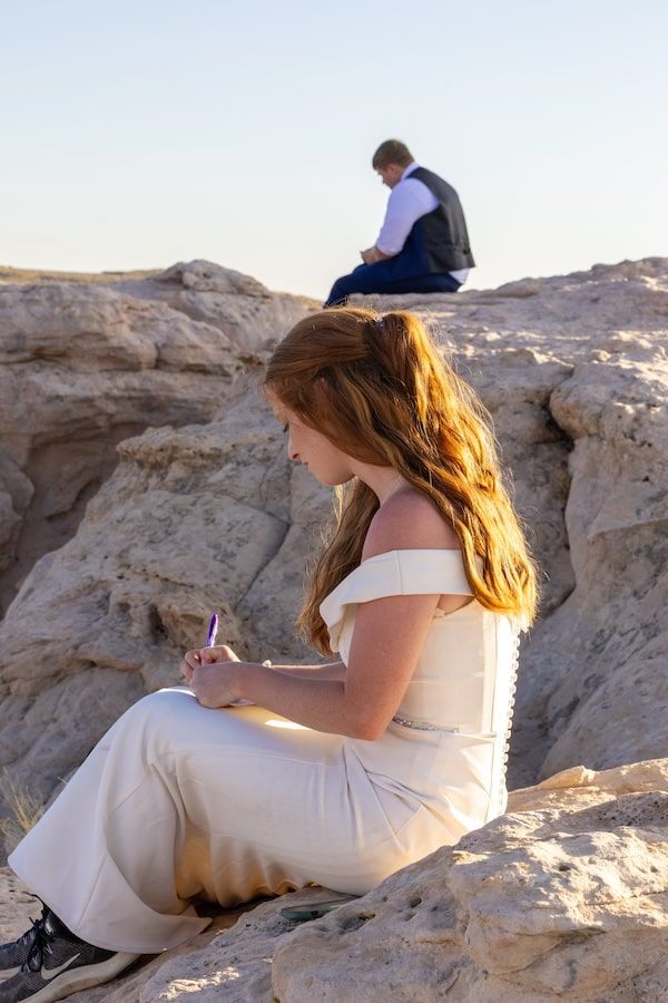 A woman in a flowing white dress sits on rocky terrain during golden hour while taking photographs.