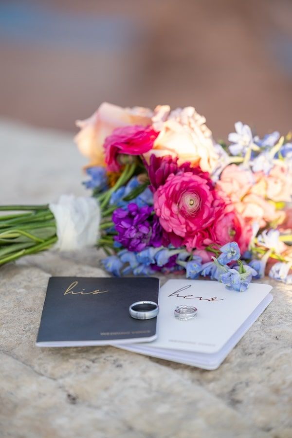 Two wedding rings sit beside black and white books with pink and purple floral arrangements on a textured stone surface.