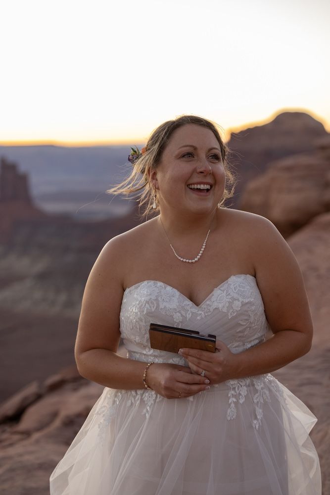 A bride in a white strapless wedding dress laughs joyfully against a scenic desert landscape at sunset.