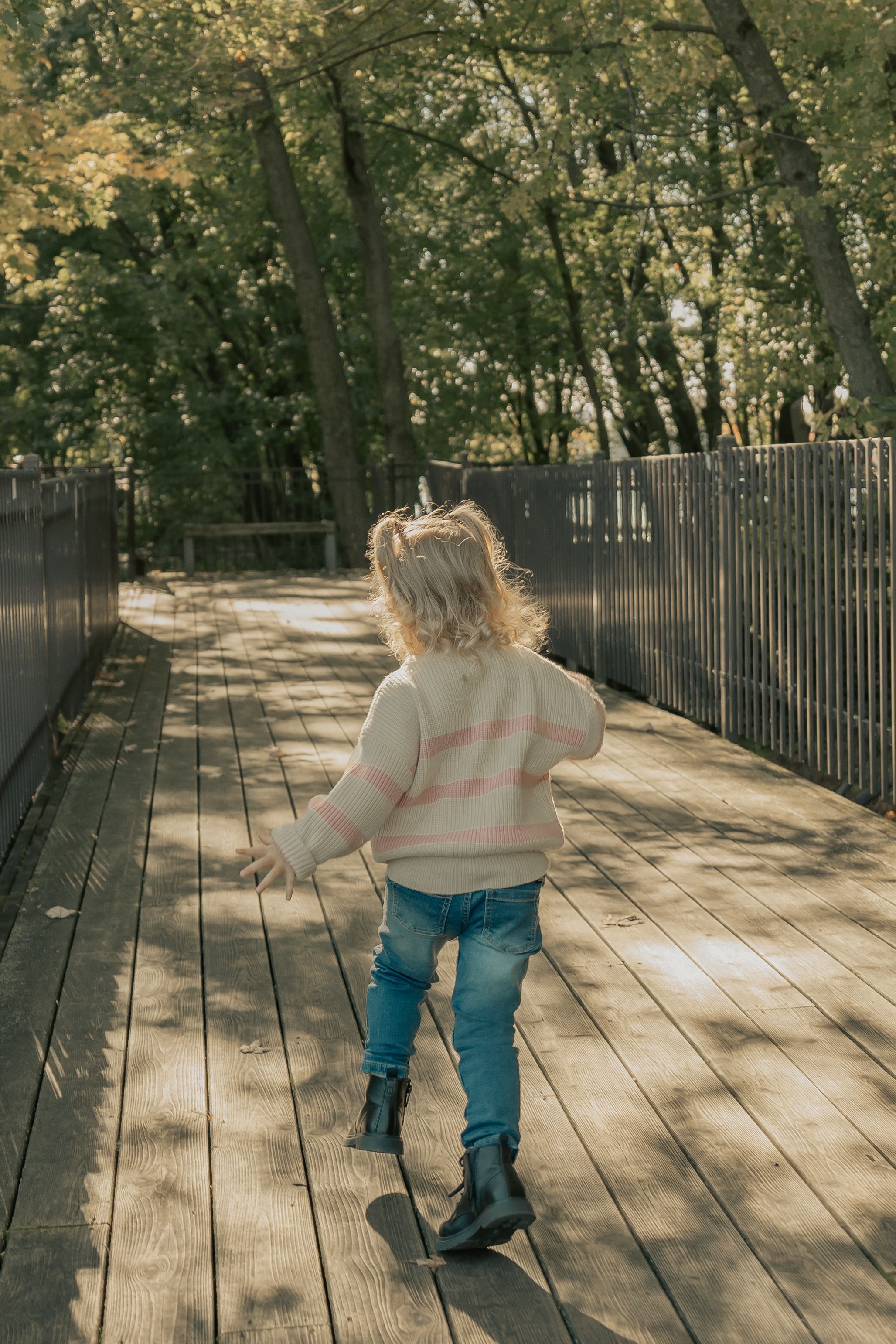 A sequence of someone walking on a wooden boardwalk path through a tree-lined park on a sunny autumn day.