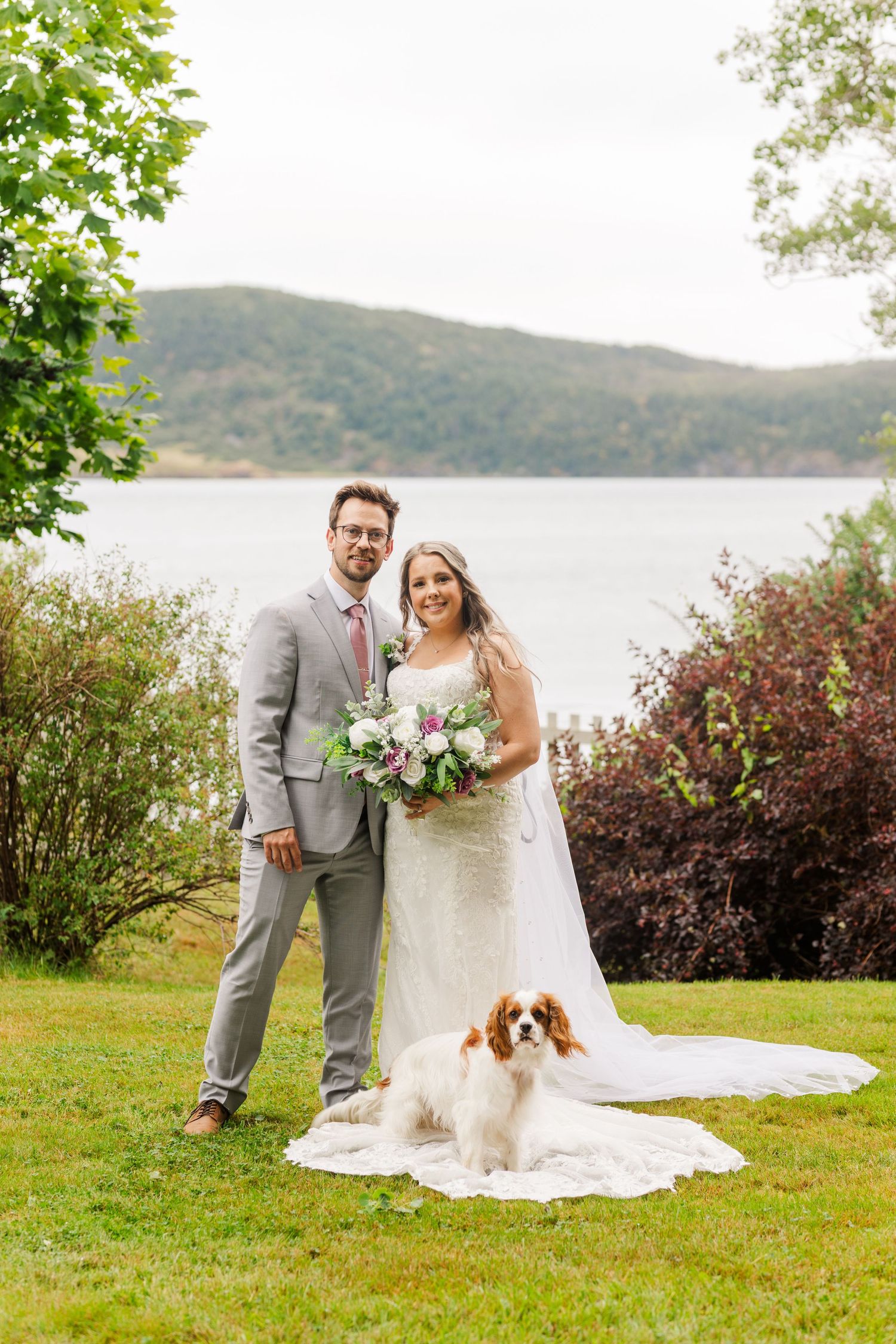 Beautiful lakeside outdoor wedding photo with newlyweds and their Cavalier King Charles Spaniel dog on grassy shore.