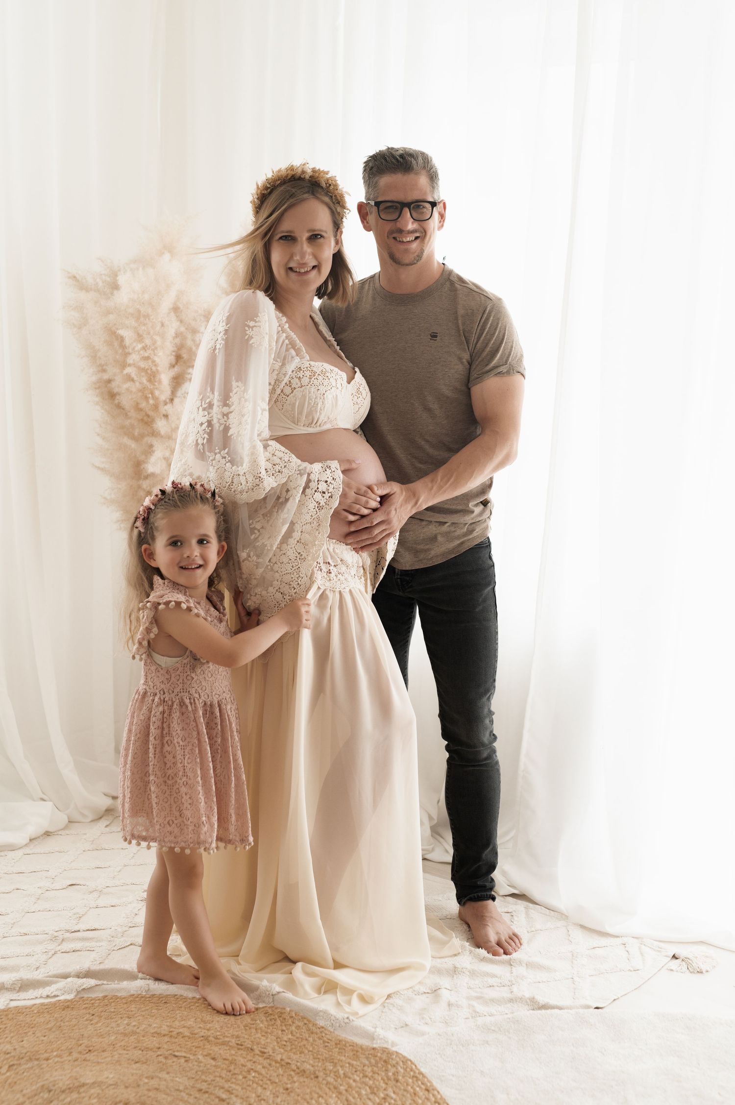 A family maternity photo featuring a lace-dressed pregnant woman, young girl in pink dress, and man against a light backdrop.