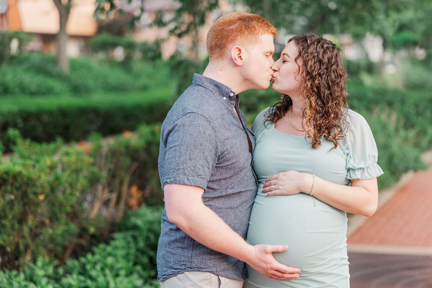 Couple sharing intimate moment during outdoor maternity photoshoot in a garden setting.