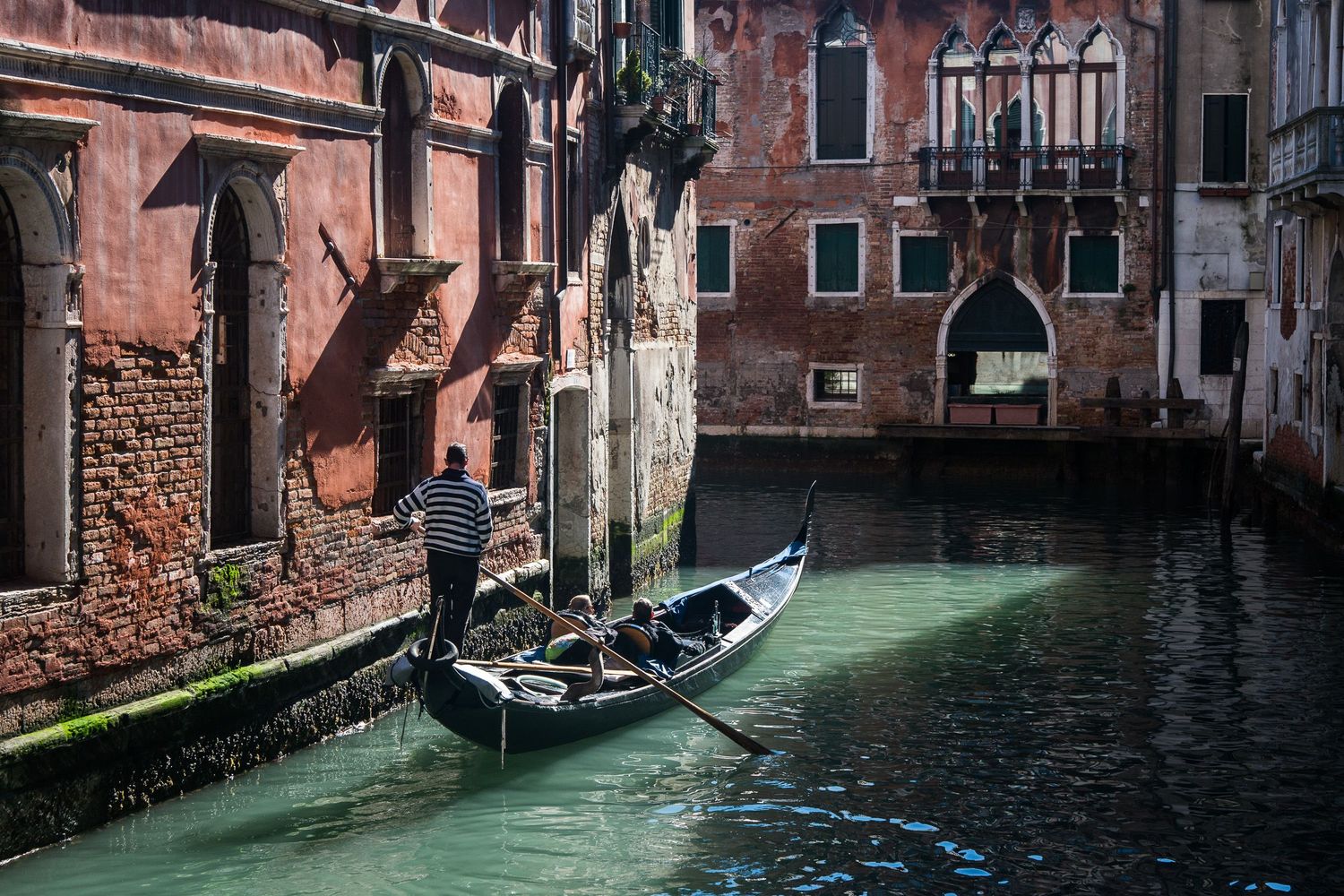 A panoramic view shows traditional gondolas and colorful buildings along a narrow Venetian canal in late afternoon light.