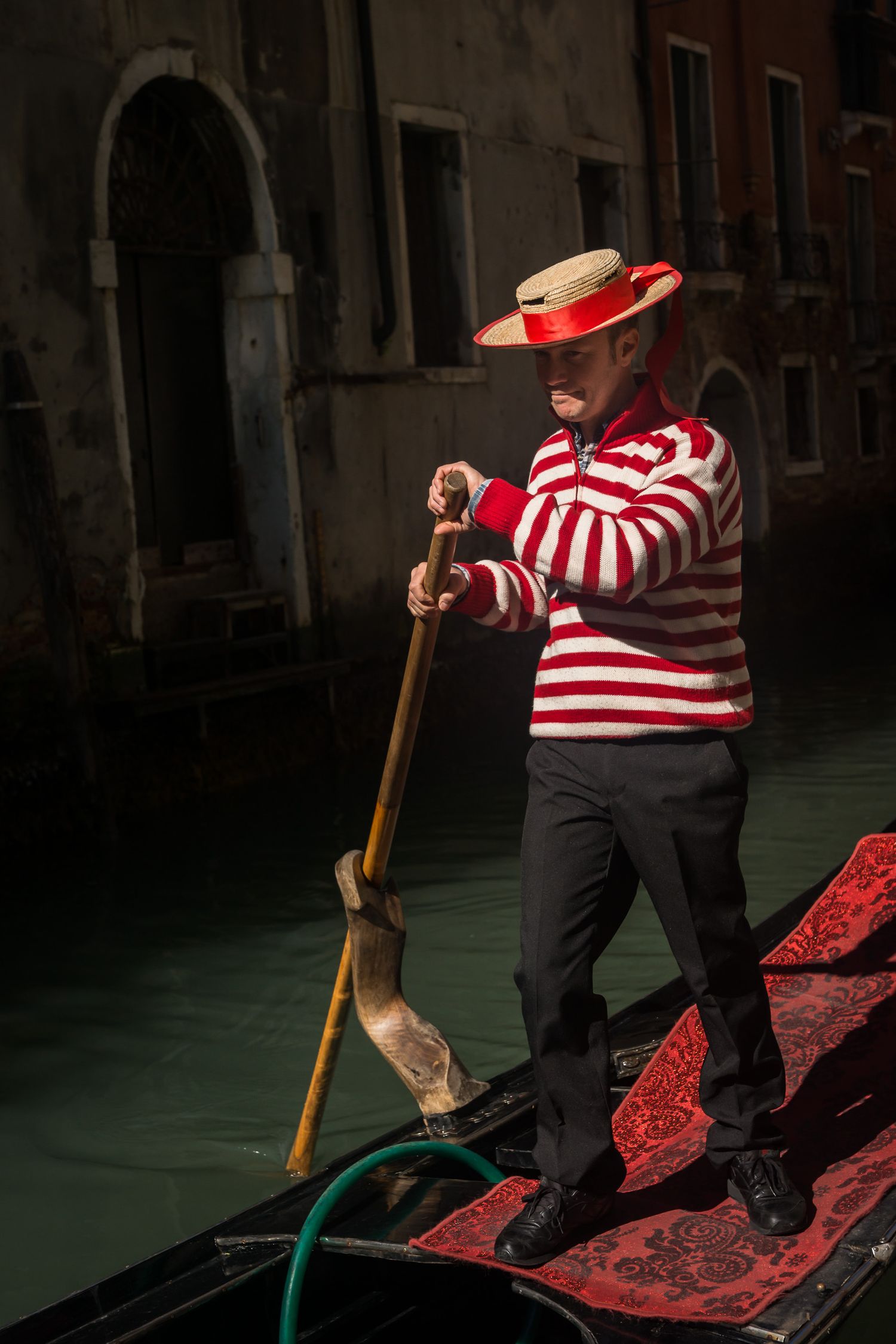 A gondolier in traditional red and white striped shirt steers a boat through Venice's dark green canal waters.
