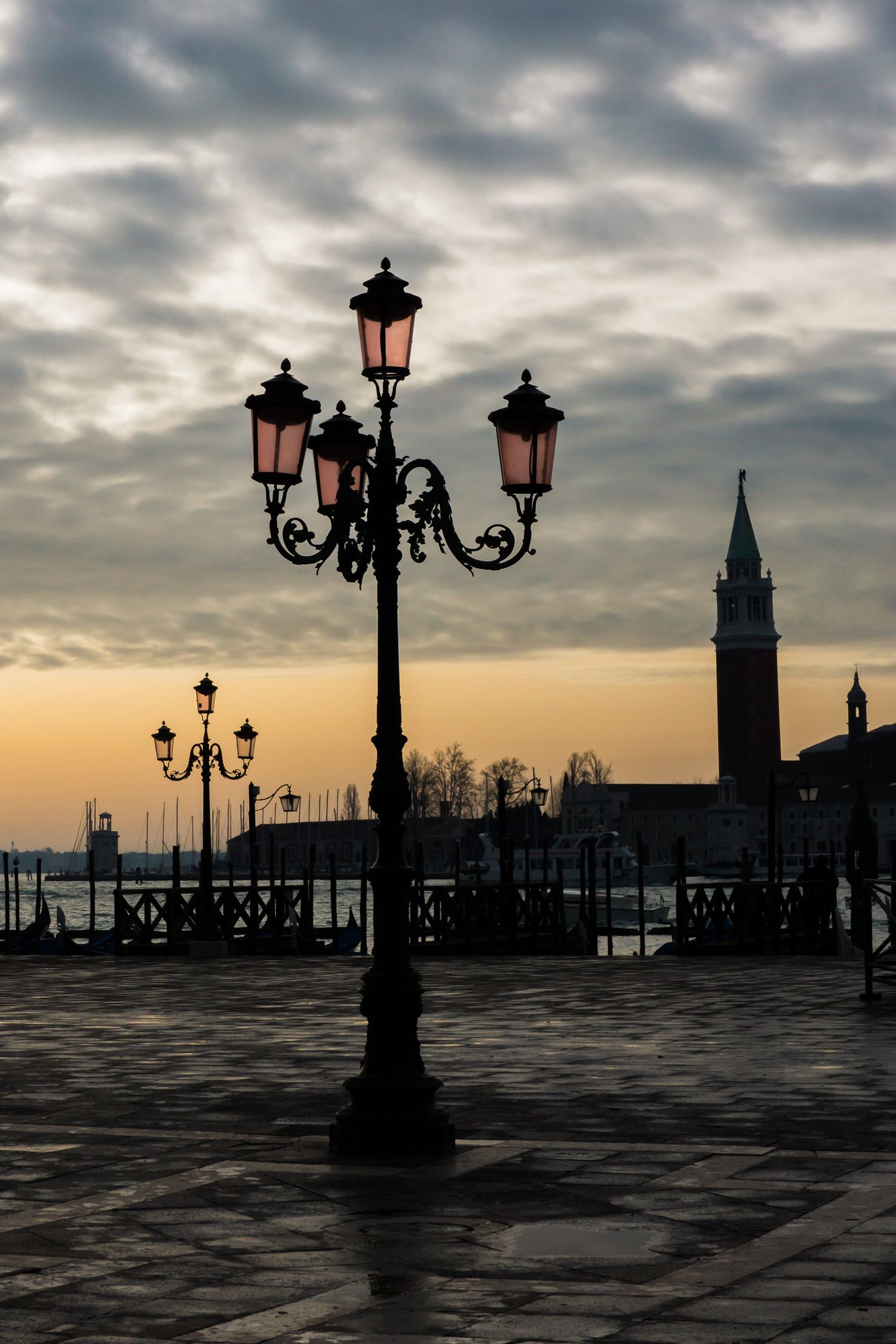Silhouetted street lamps and buildings in Venice at sunset against a moody clouded sky over the Grand Canal waterfront.
