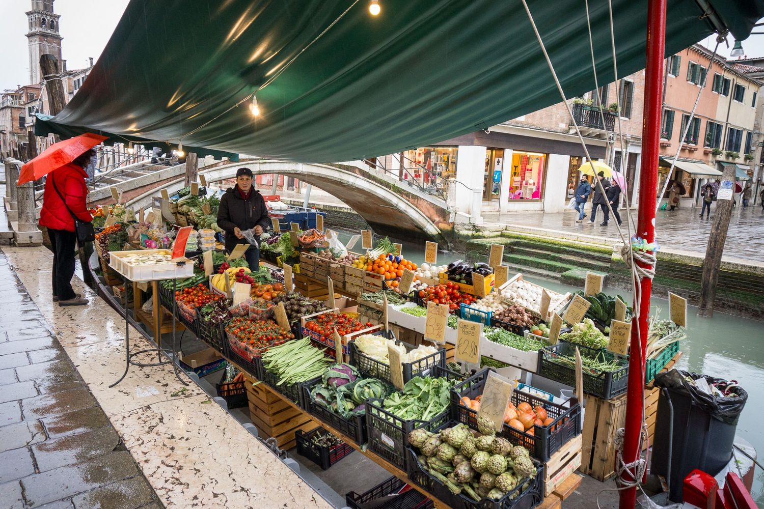 A floating market stall in Venice displays fresh produce and vegetables under a green canopy along a canal.