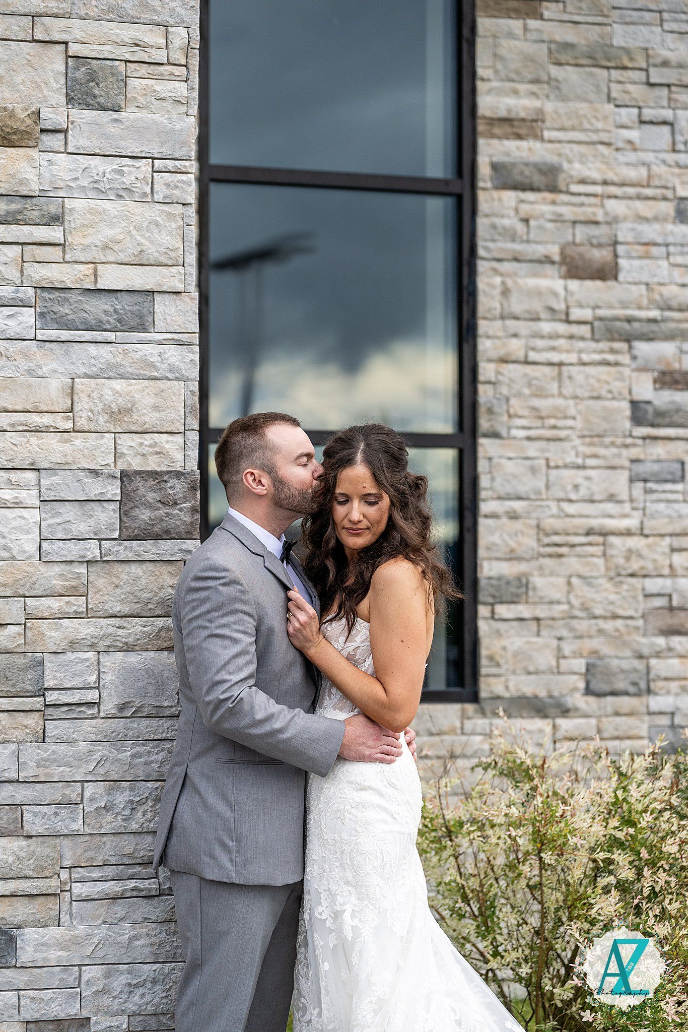 A romantic moment shared between a bride in white lace gown and groom in gray suit against a stone wall.
