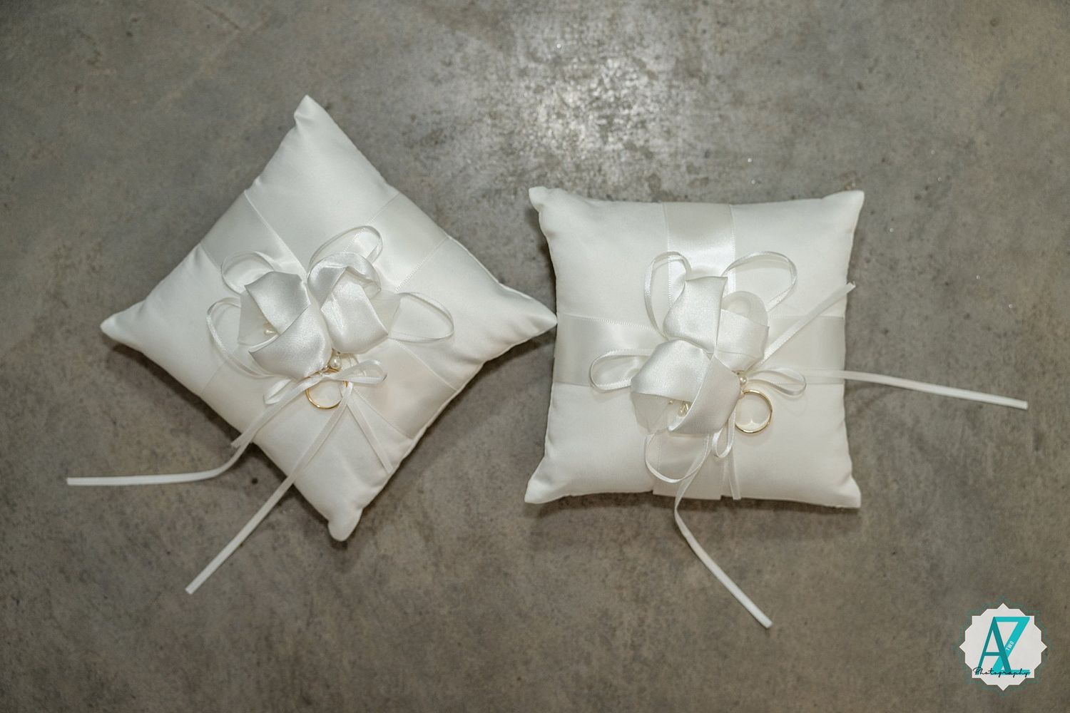 A series of white silk ring bearer pillows with ribbons and bows arranged in a progression on a concrete surface.