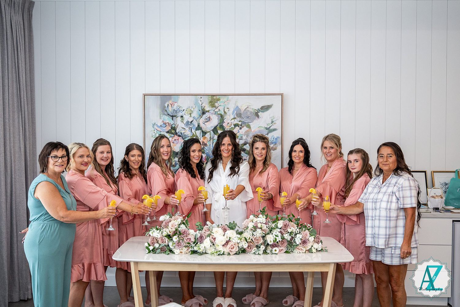A group of women in coordinated pink and teal dresses stand together in a bright room with floral arrangements.