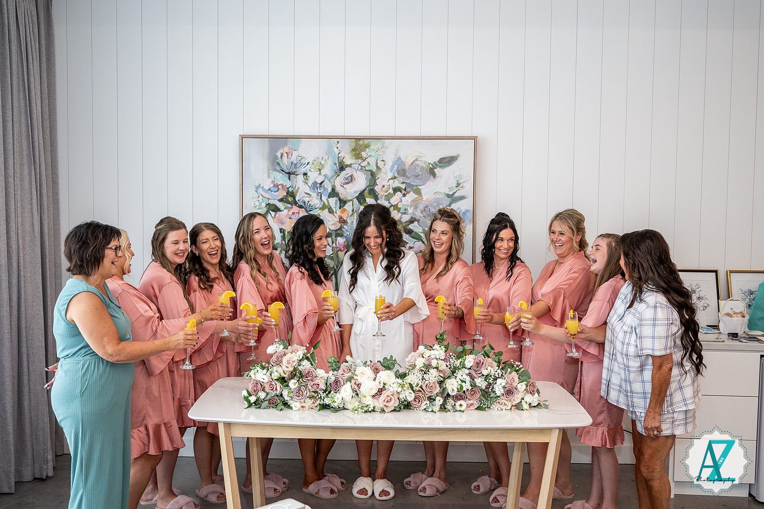 Bridesmaids in pink robes gathered around a white table with floral arrangements.