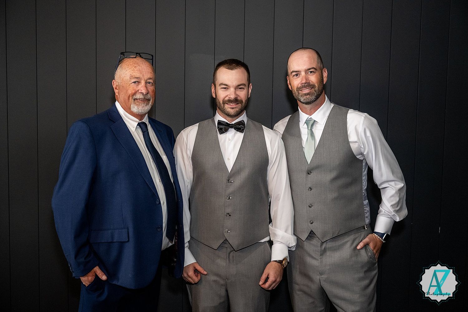 Three people in formal attire with suits and vests pose together at an event.