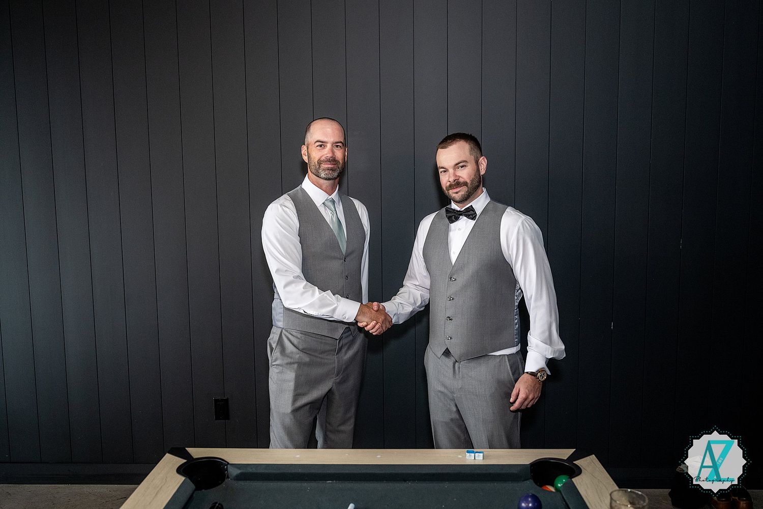 Two grooms in stylish gray suits and bow ties celebrate their wedding with cake near a rustic black wall.