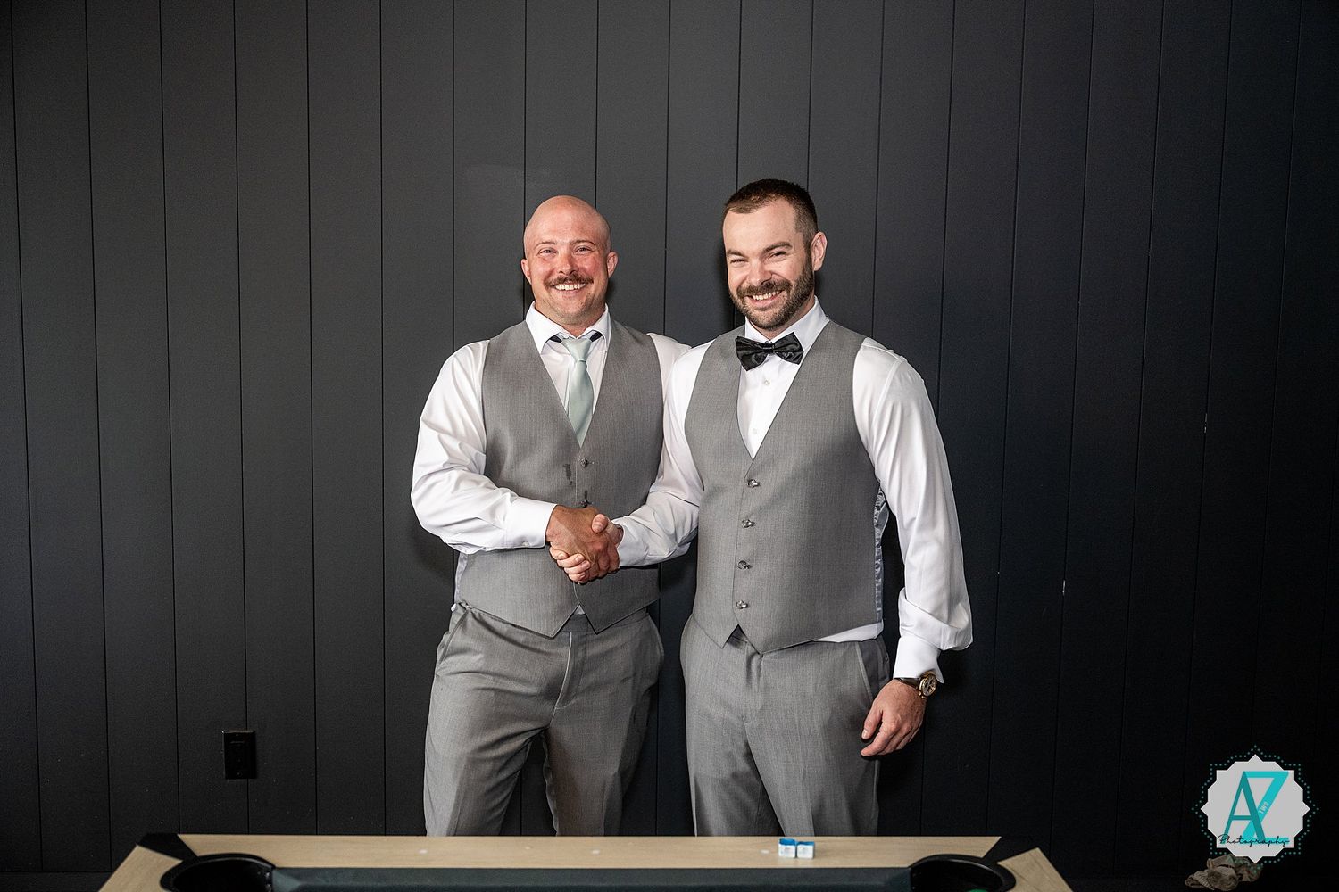 Two people in formal gray suits and vests share joyful moments against a dark wooden wall background.