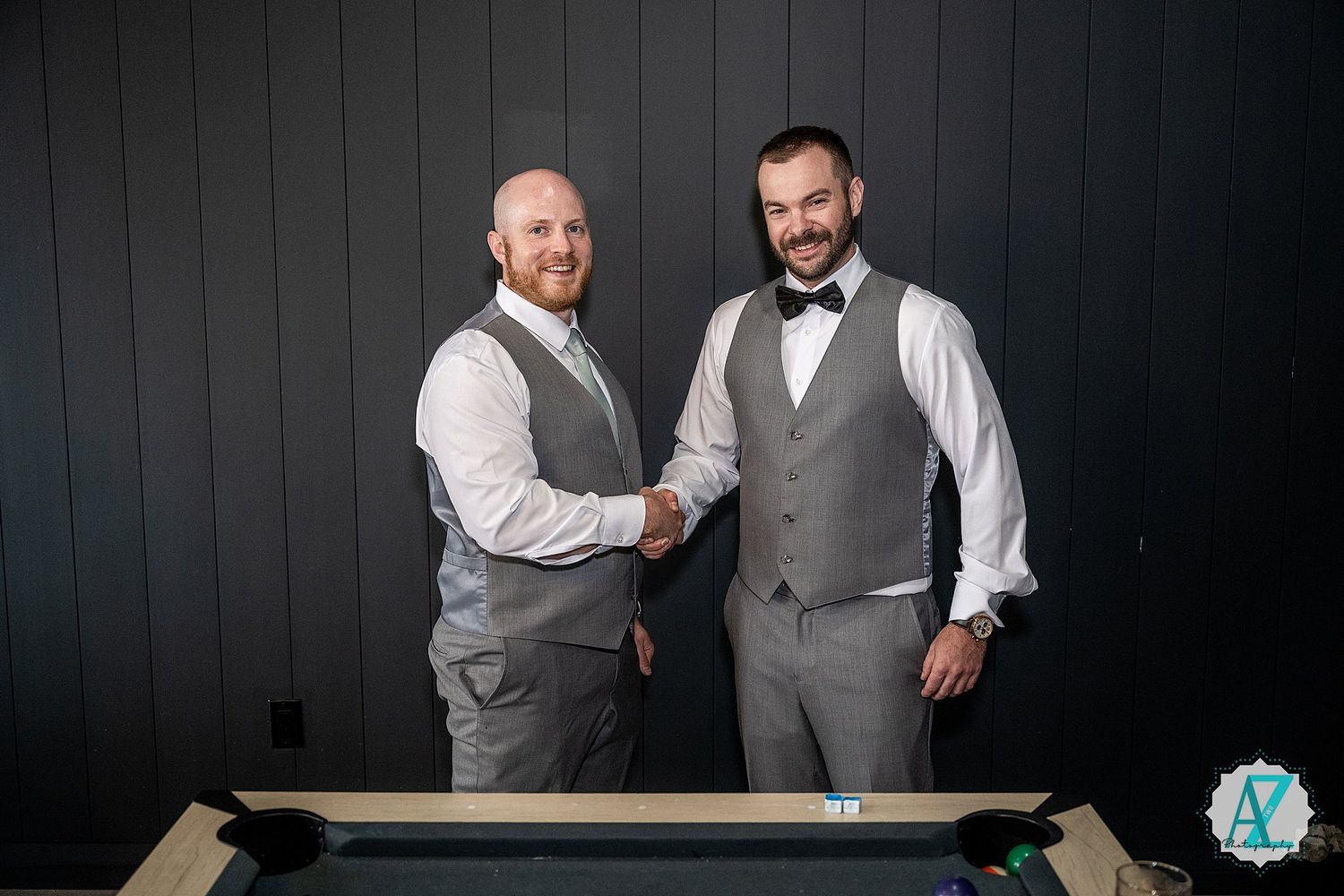 Three suited groomsmen pose playfully against a dark wall in matching gray vests and white shirts.