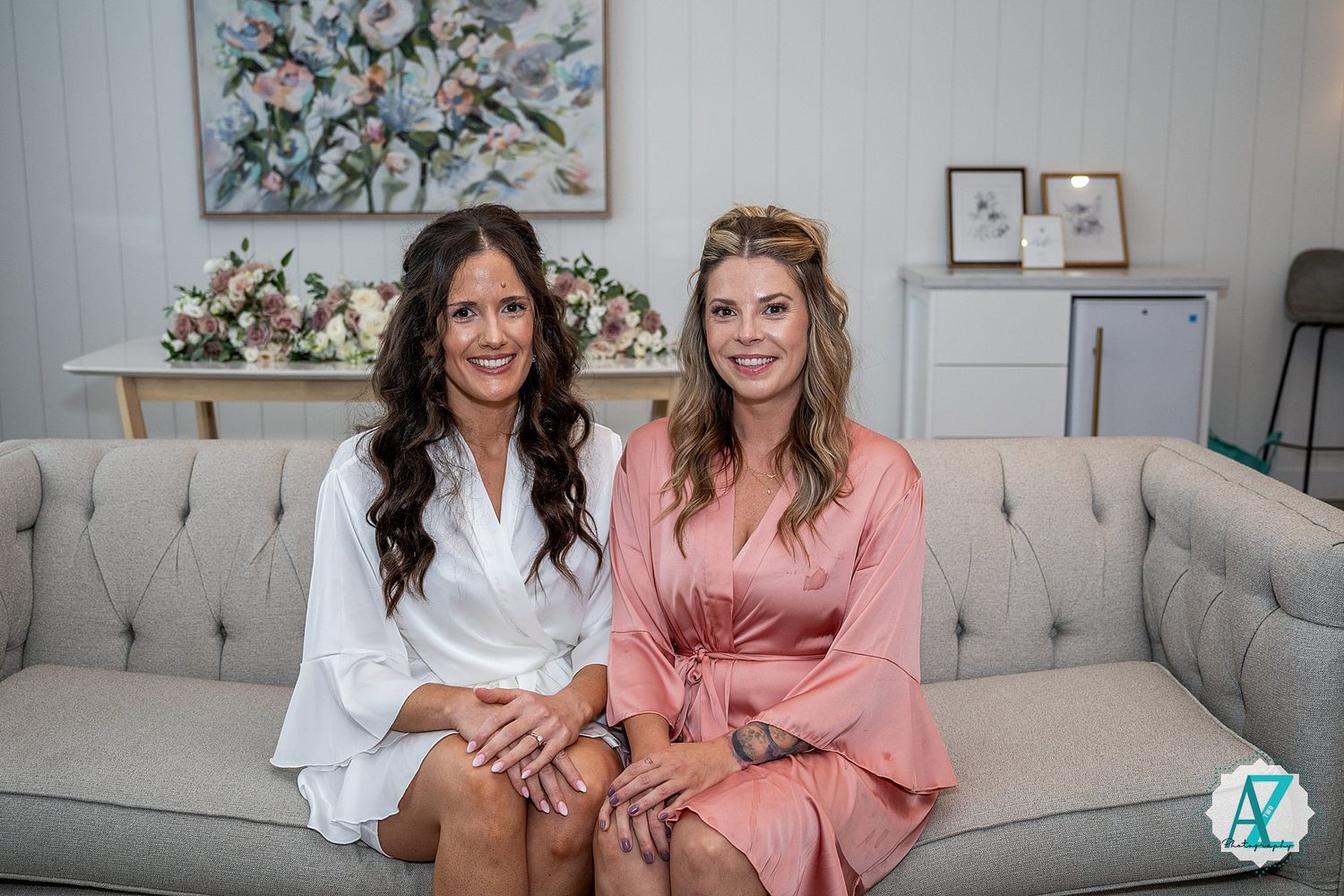Two people sitting on a grey couch pose and laugh while holding phones in a modern living room with floral artwork.