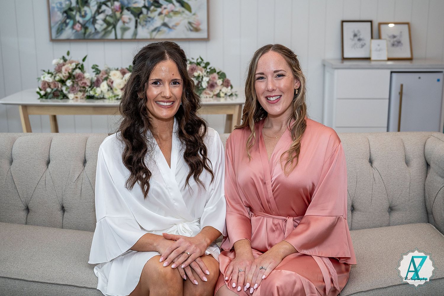 Two women sit together on a grey couch while smiling and laughing in a bright, modern living room.