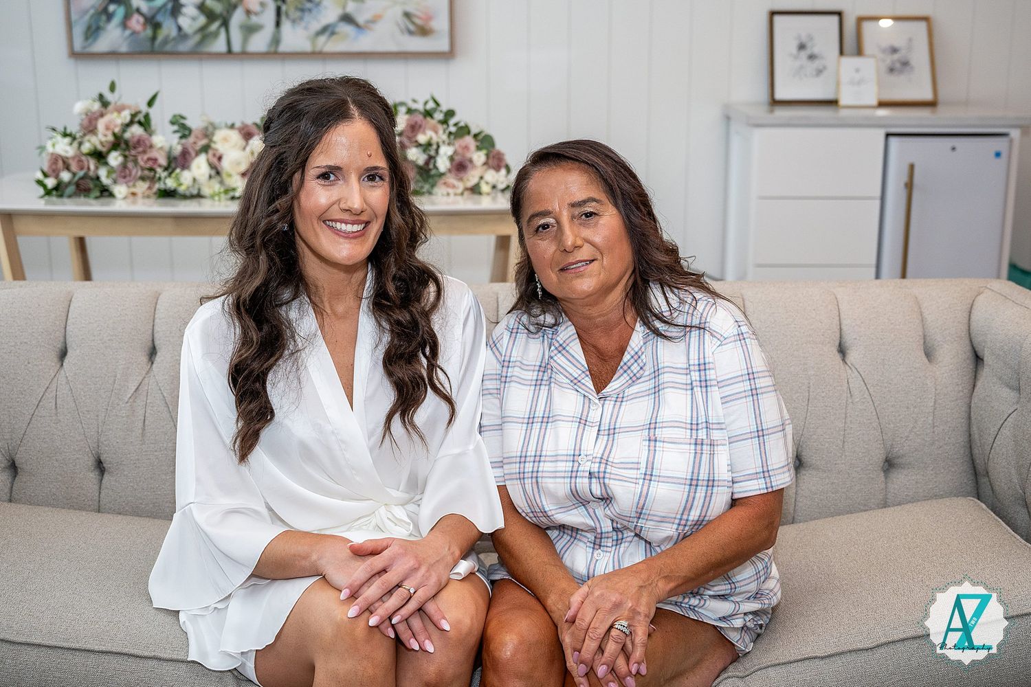Two women pose together on a light gray couch with floral artwork and white walls in the background.