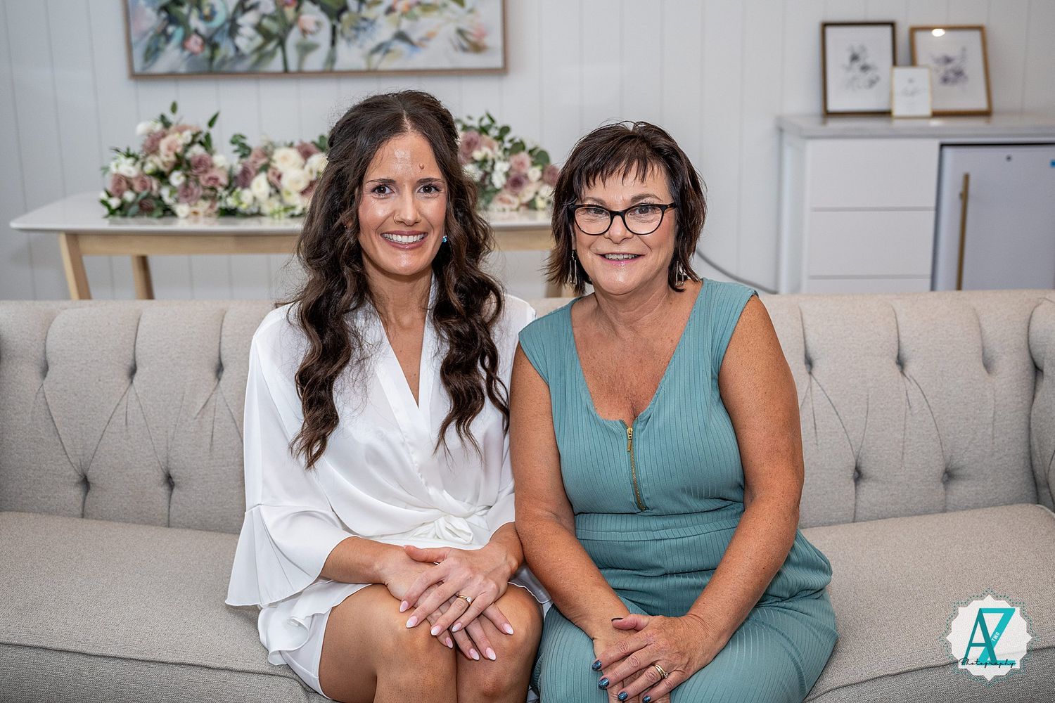 Two women share casual moments on a beige sofa with decorative wall art behind them.