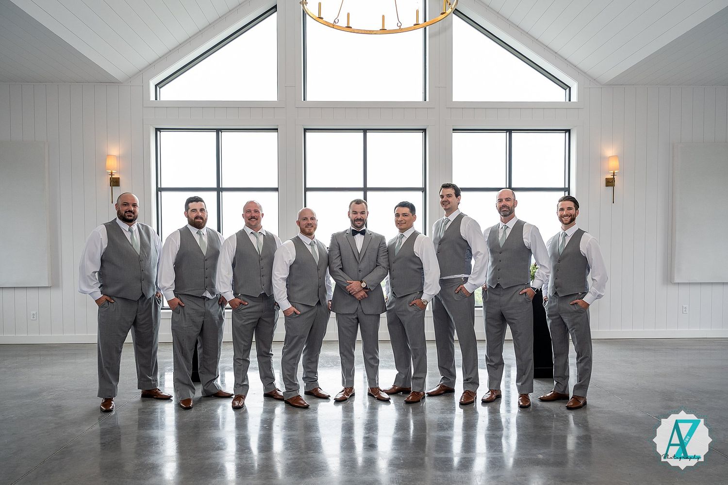 Wedding party members in gray suits stand in line against white walls and windows in bright indoor venue.