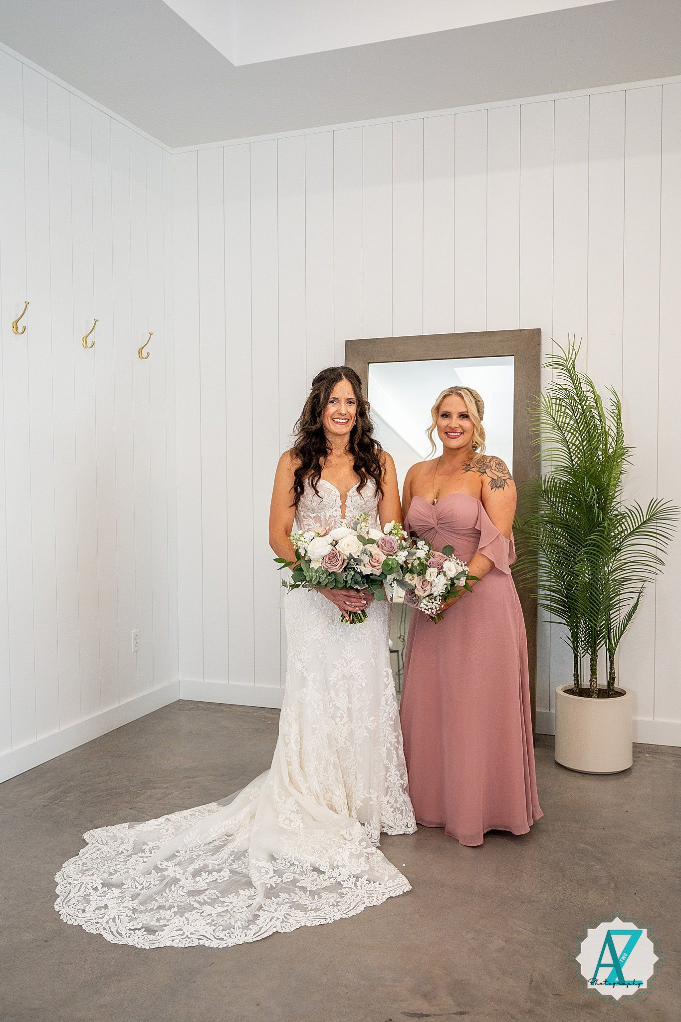 Bride in lace wedding gown and bridesmaid in dusty rose dress pose and smile together in white-walled venue.