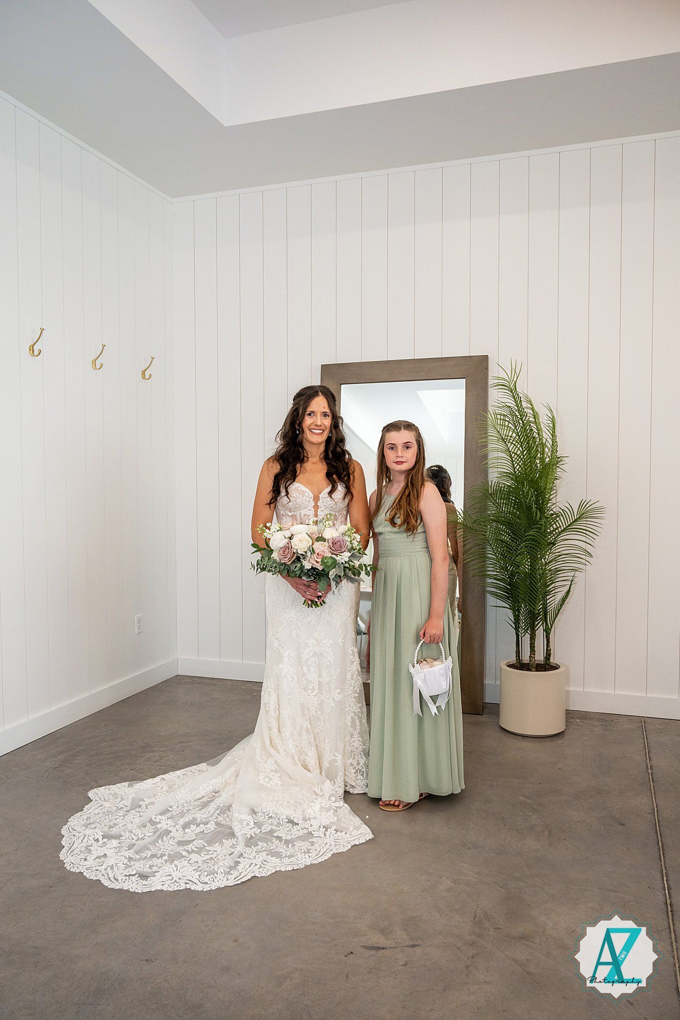 Two people standing together in an elegant setting with one wearing a white lace wedding dress and another in a sage green gown.