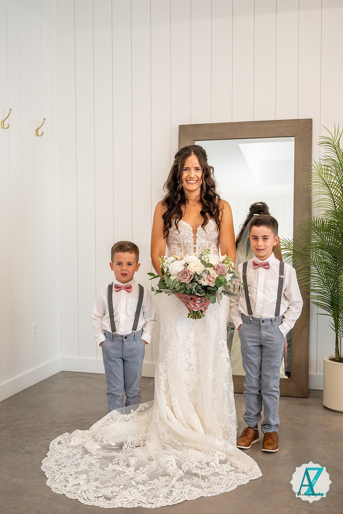 A bride in a lace wedding dress poses with young ring bearers wearing suspenders and bow ties in a studio setting.