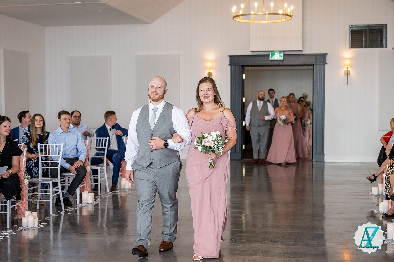 Wedding party members in gray suits and pink dresses walk down a bright white aisle during a reception ceremony.