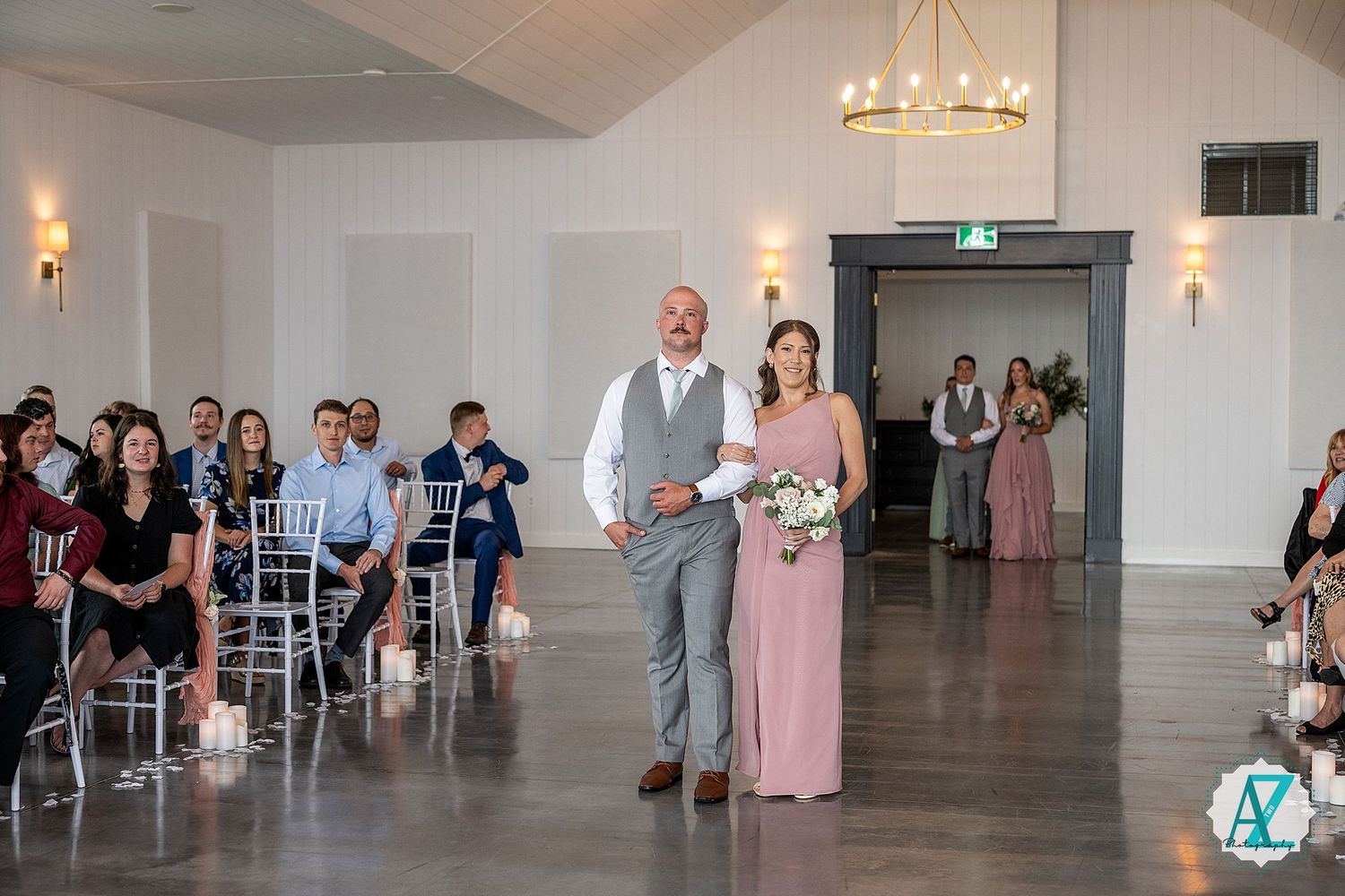 Wedding party members dance in pairs across a bright, elegant white reception hall during a formal celebration.