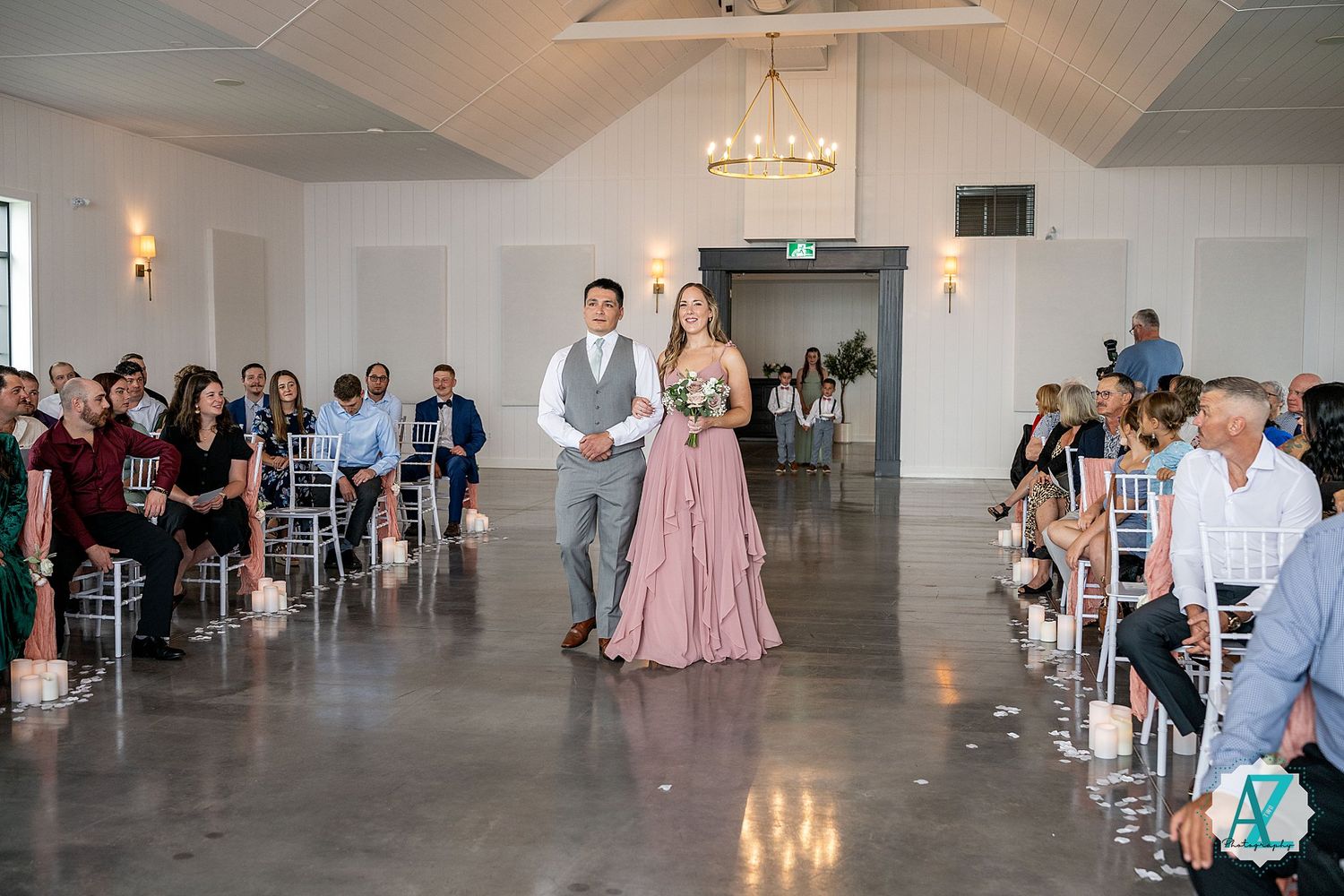 Wedding guests gather in an elegant white venue with chandeliers and gray flooring for a celebration.