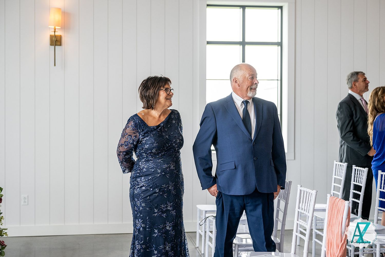 Wedding guests mingle in a bright white venue with windows and white chairs near wall-mounted sconces.