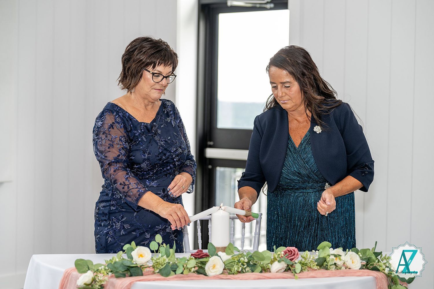 Two women in navy blue dresses arrange flowers and decorations on a table at an event.
