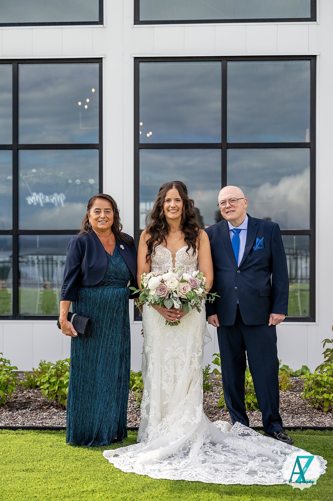 Wedding party poses outdoors against modern white building with floor-to-ceiling windows on cloudy day.