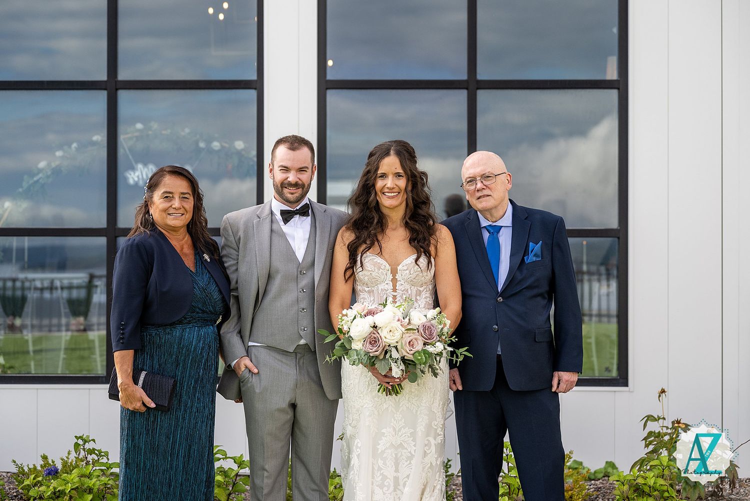 Wedding party poses in front of modern white building with large windows and cloudy sky backdrop.