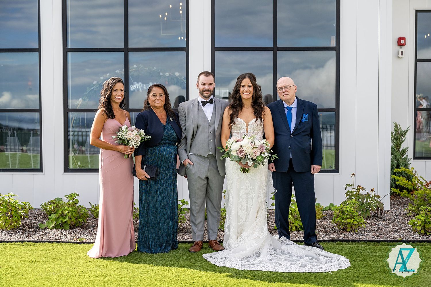 Wedding party poses outdoors at a scenic venue with modern glass windows and lush landscaping in the background.
