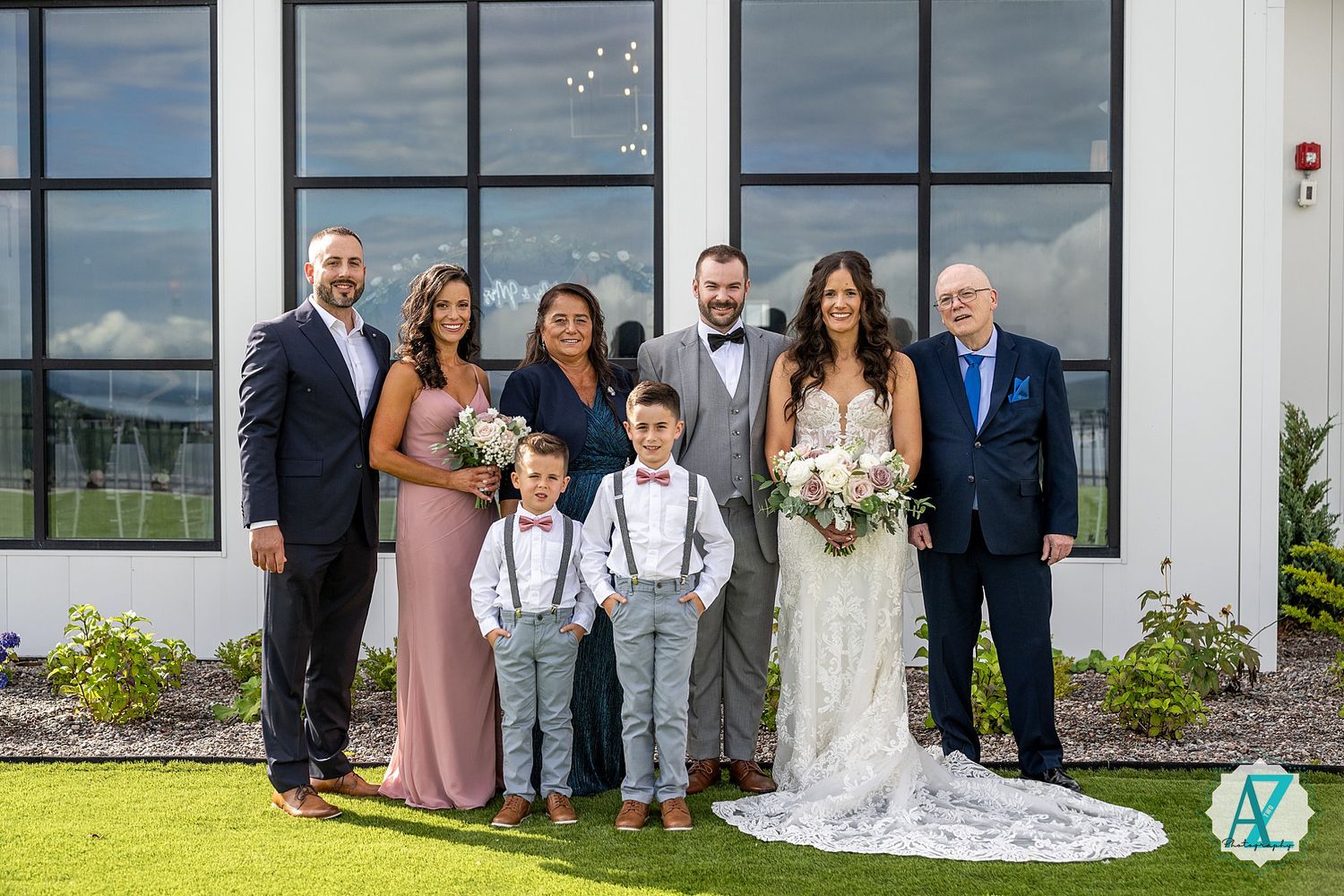 Wedding group photo outside a contemporary white building with dark windows and landscaping.